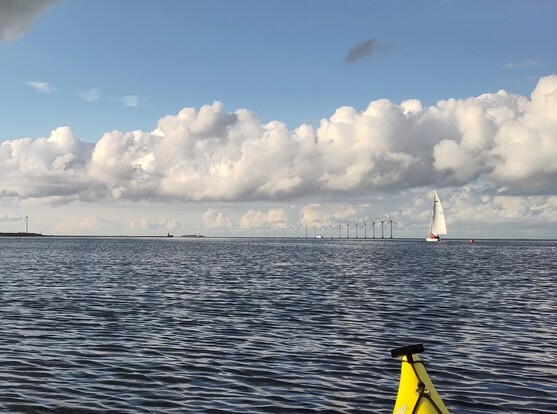 Yellow kayak tip on calm blue sea with blue sky and fluffy clouds overhead. In the far distance a line of wind turbines and a white sailing boat