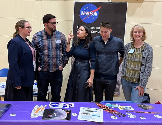 Four scientists surround Congresswoman AOC as she talks about stem education. NASA poster in the background and swag in the foreground. 