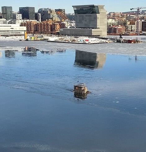 A small square boat looking like a shed, floats on the water in front of modern high buildings under a blue sky reflected in the water 