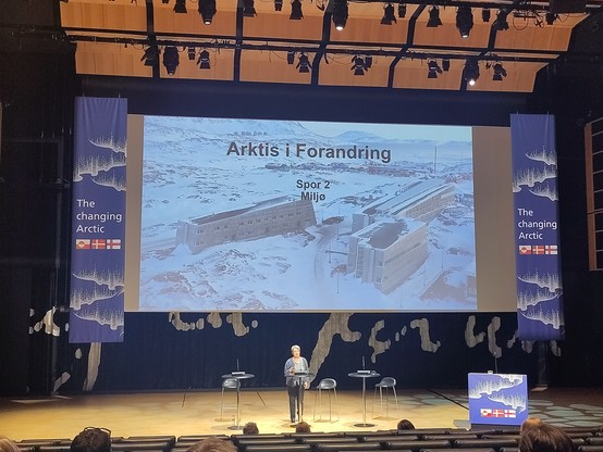 Woman with short grey hair standing on a stage in front of title slide reading Arktis i forandring with a picture of a modern blocky building in an open snow covered landscape