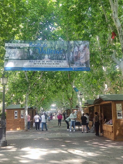 En la foto se ve una arboleda, unas casetas, gente y el cartel de la Feria del libro de Vallecas 