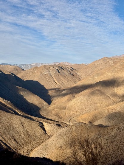 A dry mountain valley below Cerro Pachón.  Dry desert moutons are seen with a. blue partly cloudy sky above. Higher partly snow covered mountains are seen in the distance.