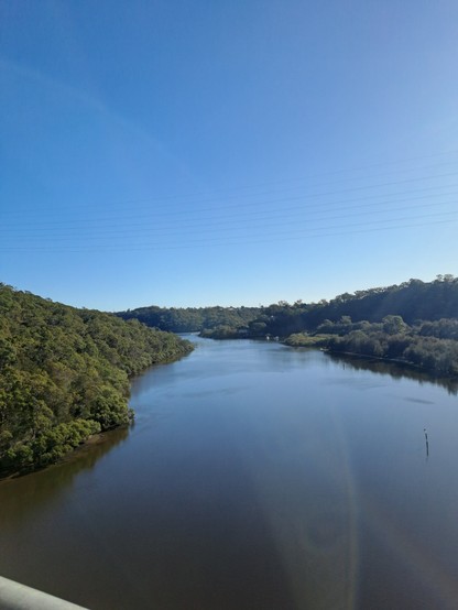 A view of a river from a high bridge. The river is wide, with green treed banks on either side. The bright blue of the cloudless sky reflects on  the brownish water.