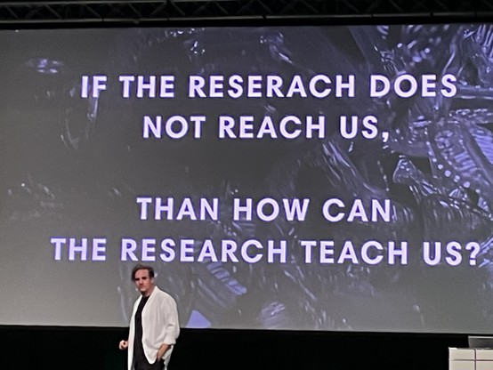 The presenter in white jacket and black clothes underneat a big b/w slide reading 
“IF THE RESERACH DOES NOT REACH US, 

THAN HOW CAN THE RESEARCH TEACH US?”