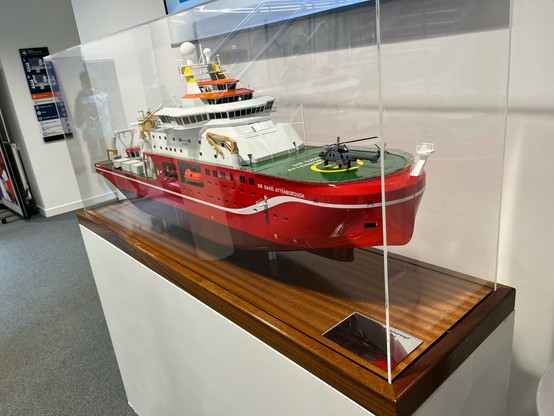 ~1:100 scale model of a 189m ship with an orange hull, white research decks and green bow with a helicopter on top. “RSS Sir David Attenborough”