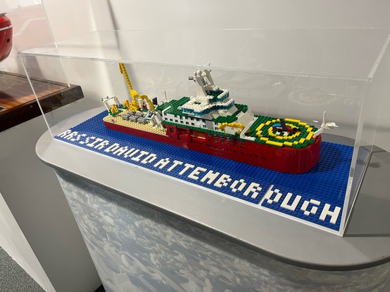 In LEGOS ~1:200 scale model of a 189m ship with an orange hull, white research decks and green bow with a helicopter on top. “RSS Sir David Attenborough”