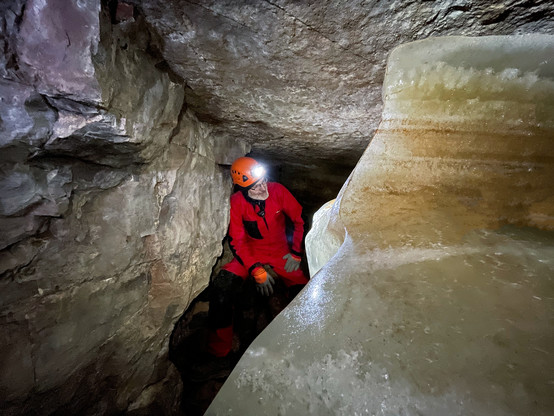 A person in a red caving suit crouching before a wall of layered ice.  He is wearing an orange helmet with a bright headlamp.
