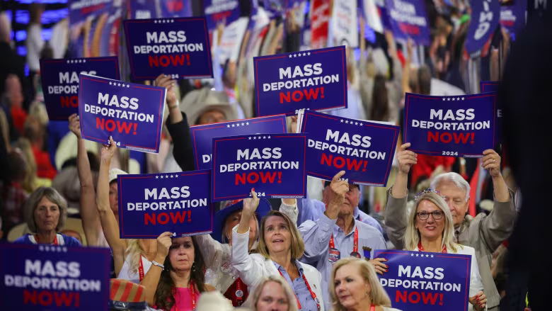 White women and a couple of older white guys hold up official-looking red, white, and blue signs reading "Mass Deportation Now" at the RNC.