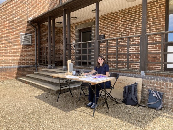 Photo of a woman behind a table outdoors in front of a building, waiting to welcome participants