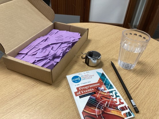 Photo of a box full of tickets that read “Tuesday dinner”, concert programme and glass of water on a small
Table
