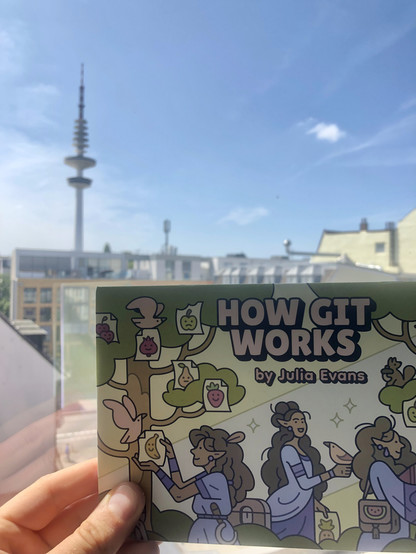 A small booklet / zine entitled “How Git Works” by Julia Evans is held in front of a window with a view of the radio telecommunication tower in Hamburg, Germany against a blue sky. 