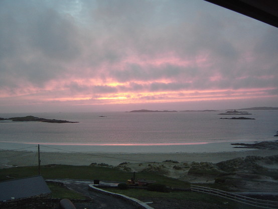 Evening sun at the coast. Small road and pasture in the very front; silvery to purple (reflections of the sun) sea; dark grey clouds with bright orange to purple streaks and beams of the evening sun.