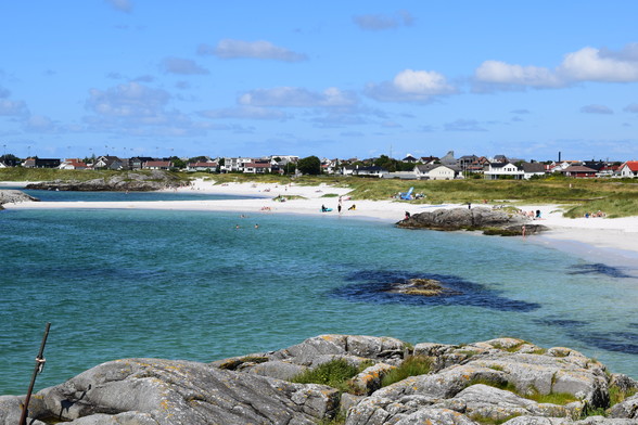 A photograph of a sandy beach and azure waters, taken from a small rock outcropping.