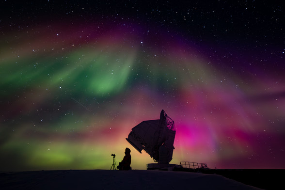 a person in silhouette taking a picture of the sky with a telescope dish in the background. The night sky is filled with green and pink auroras.