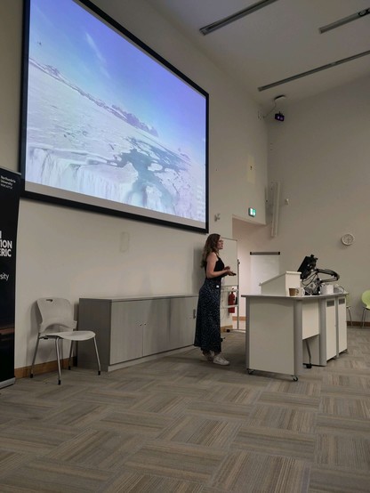 Woman in a long dress standing by a computer and in front of a screen where a film shows a waterfall off an ice shelf edge
